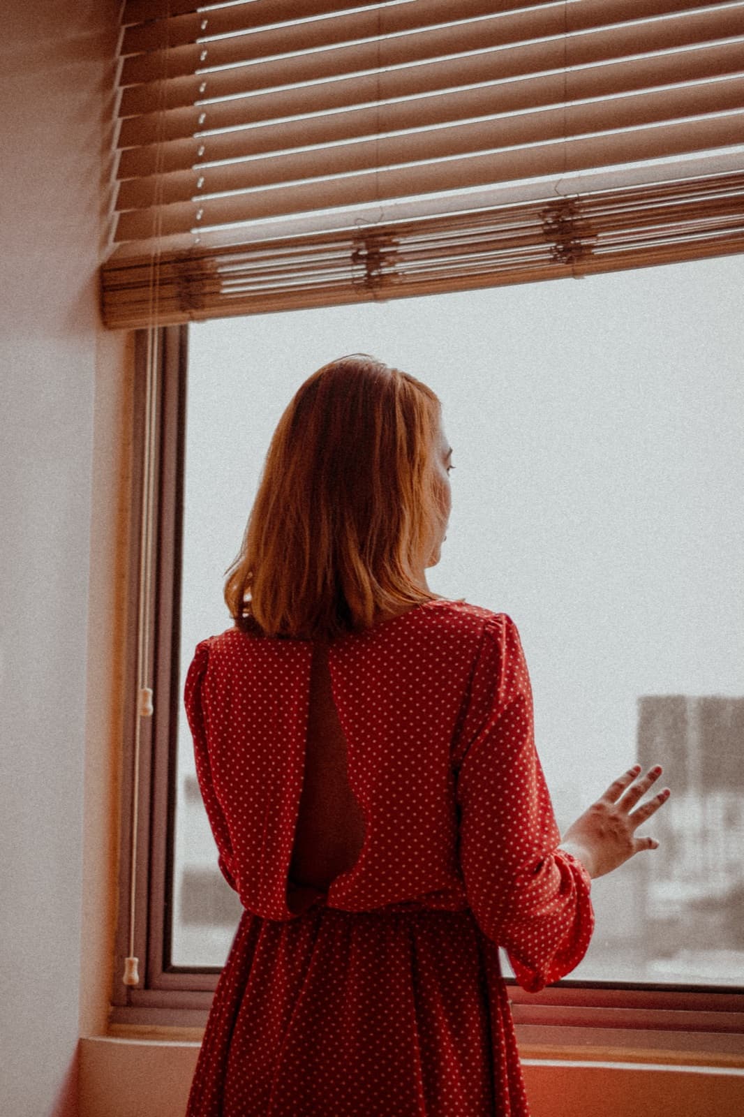 A parent standing at a window in an ordinary room, looking out, mid-thought.