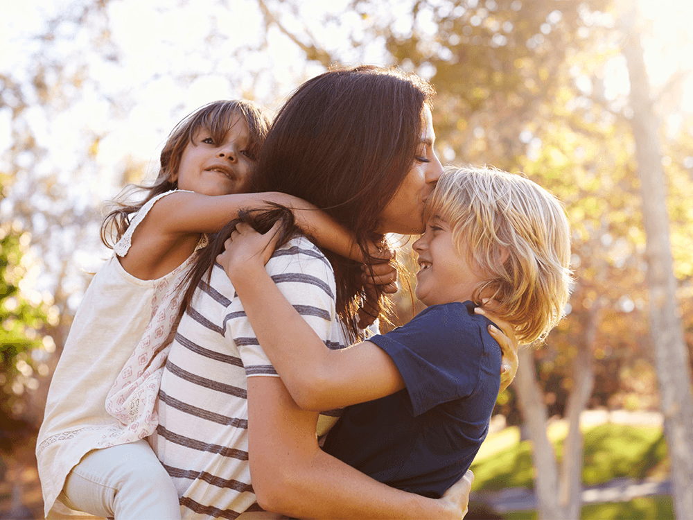 A parent outdoors being hugged by two young children.